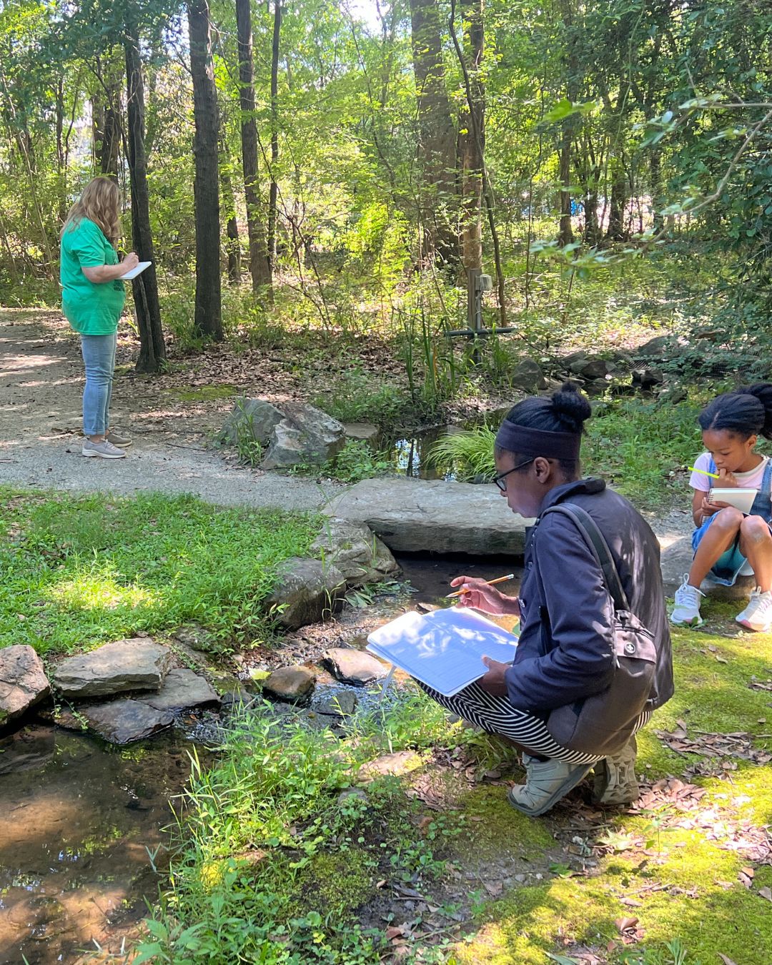 People outdoors in a wooded area beside a small stream. One person crouches near the water holding a sketchbook and pencil, another kneels nearby with a notebook, and a third stands on a nearby path holding paper. Trees, rocks, and greenery surround the scene.