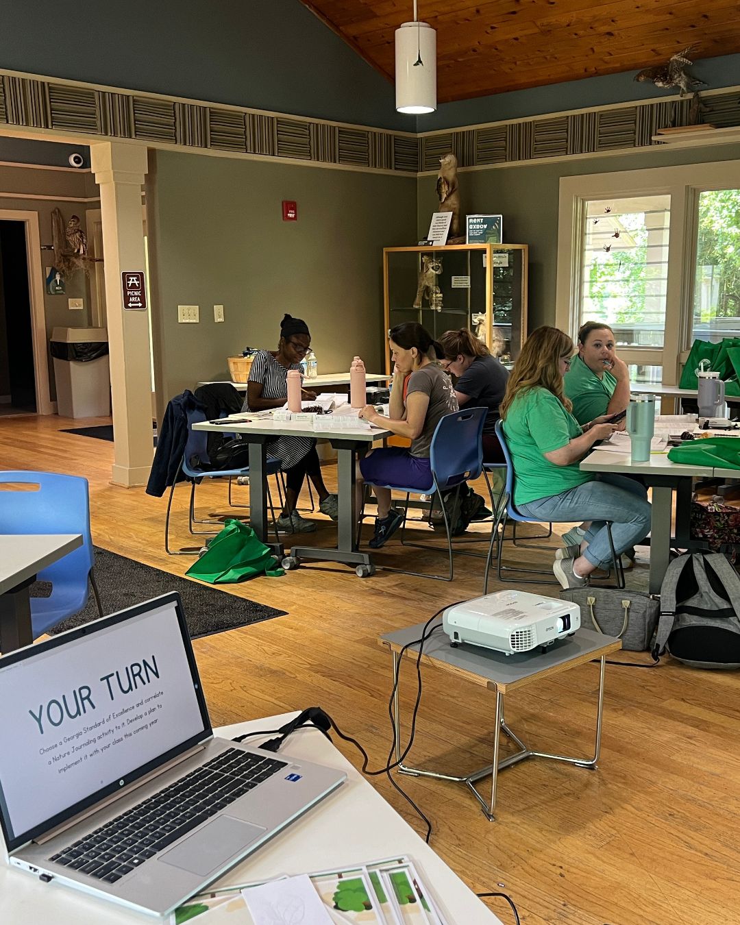 Indoor classroom setting with several people seated at tables writing and reading. In the foreground, a laptop on a table displays the text ‘YOUR TURN,’ connected to a projector on the floor. Notebooks, water bottles, and bags are visible, with display cases and windows in the background.