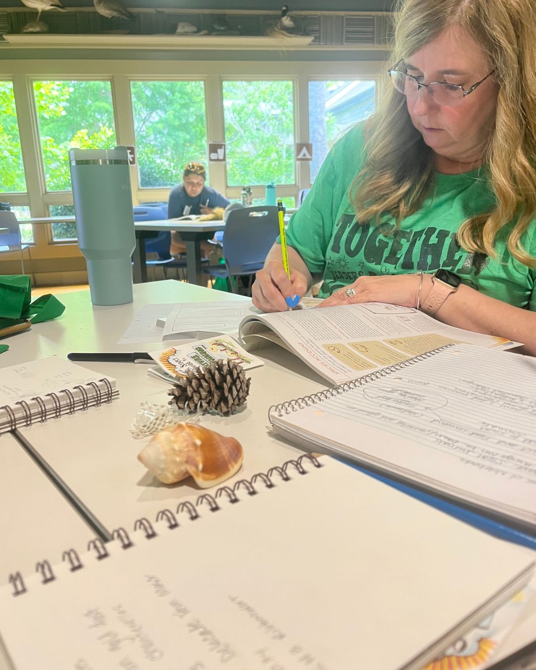 Person seated at a classroom table writing in an open workbook with a pencil. Notebooks, a pinecone, a shell, and a reusable tumbler are on the table, with other people working at tables in the background near windows.
