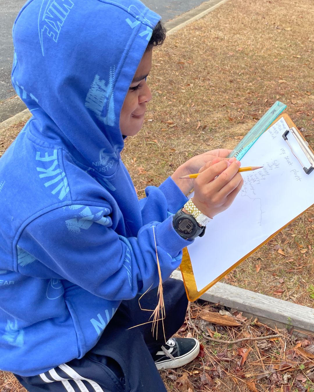 Person seated outdoors beside a curb, wearing a blue hooded sweatshirt, holding a clipboard and pencil while writing or sketching on paper, with a ruler resting on the clipboard.